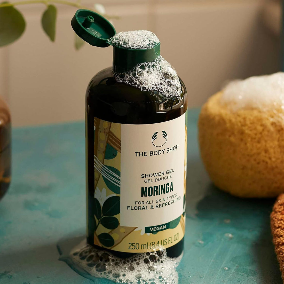The Body Shop Moringa shower gel bottle with a sponge on a blue surface